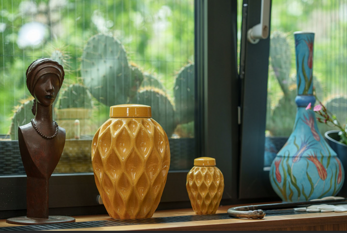 Golden Echo 3D printed ceramic urn with flowing lattice pattern on a windowsill beside a bust and blue vase