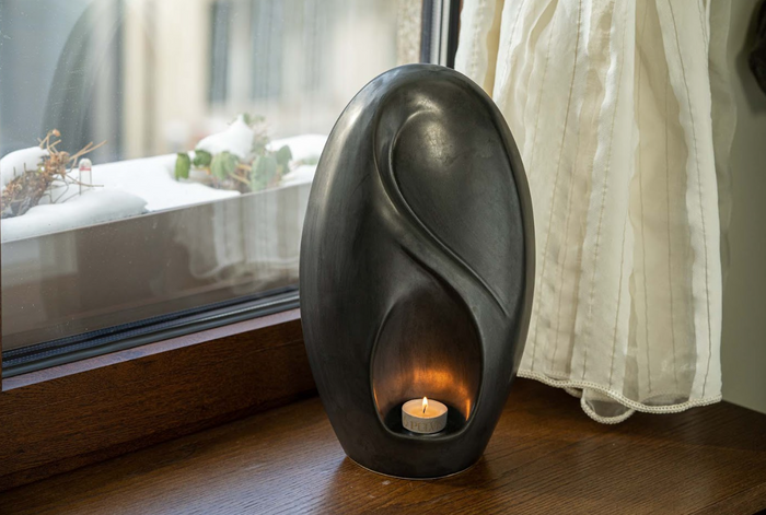Eternity Ceramic Urn in dark charcoal with a sculptural curved design and a candle glow by a window.