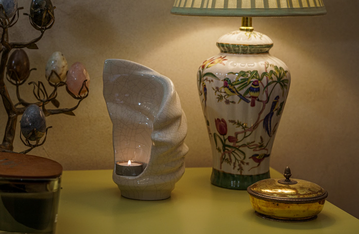White crackled ceramic candle urn with inner cavity holding a small tealight on a yellow table beside a decorative vase.