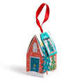 A small red cardboard house with a single gabled roof hangs from a red ribbon. Snowflake patterns twinkle across the surface, with colorful green, yellow, purple, and blue illustrations. The front has a blue door with a tiny white window above it, and two decorated Christmas trees on the side. A product tag with a Christmas tree illustration hangs one side of the roof.