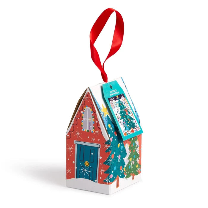 A small red cardboard house with a single gabled roof hangs from a red ribbon. Snowflake patterns twinkle across the surface, with colorful green, yellow, purple, and blue illustrations. The front has a blue door with a tiny white window above it, and two decorated Christmas trees on the side. A product tag with a Christmas tree illustration hangs one side of the roof.