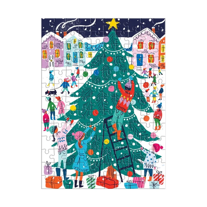 The image shows a completed puzzle from the small red cardboard house, forming a bright winter scene. Children play in the snow in front of pastel‑colored houses. In the center foreground stands a large green Christmas tree decorated with white lights and colorful ornaments. A boy is halfway up the tree on a ladder while other children decorate below, with wrapped gifts scattered across the snowy ground.